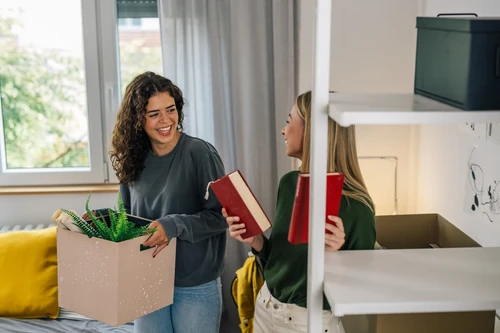 Two young women packing books and items into a cardboard box inside a bedroom